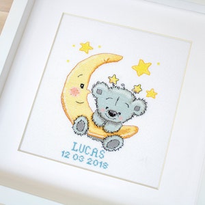 May include: Framed cross-stitch picture. A grey teddy bear smiles from a yellow crescent moon, with yellow stars around it. The name "LUCAS" and the date "12 03 2018" are stitched in blue, all within a white frame.