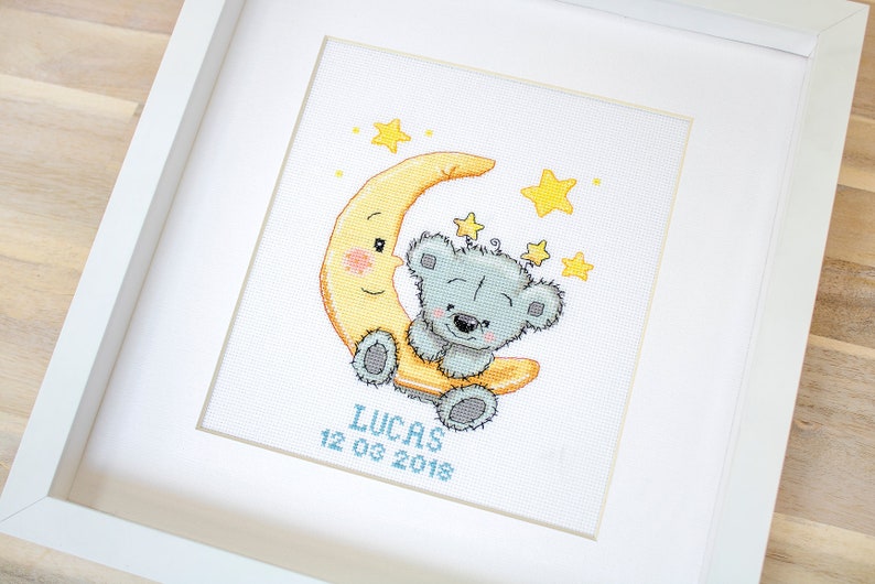 May include: Framed cross-stitch picture. A grey teddy bear smiles from a yellow crescent moon, with yellow stars around it. The name "LUCAS" and the date "12 03 2018" are stitched in blue, all within a white frame.