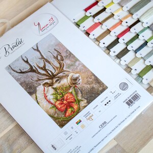 May include: A cross-stitch kit featuring a reindeer with a red bow and holly. The kit includes a white Aida cloth, floss, and a needle. The packaging is white with the text "Set de Brodat" in French.
