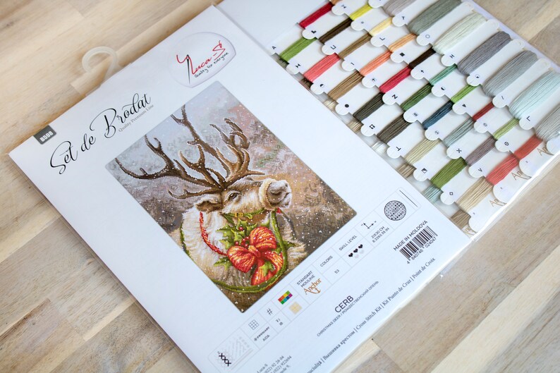 May include: A cross-stitch kit featuring a reindeer with a red bow and holly. The kit includes a white Aida cloth, floss, and a needle. The packaging is white with the text "Set de Brodat" in French.