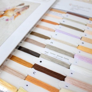 May include: A close-up of a cross-stitch kit with a variety of floss colours, including beige, brown, orange, and white. The floss is organised in labelled sections, with the text "Contour - Контур - Contorno" visible.