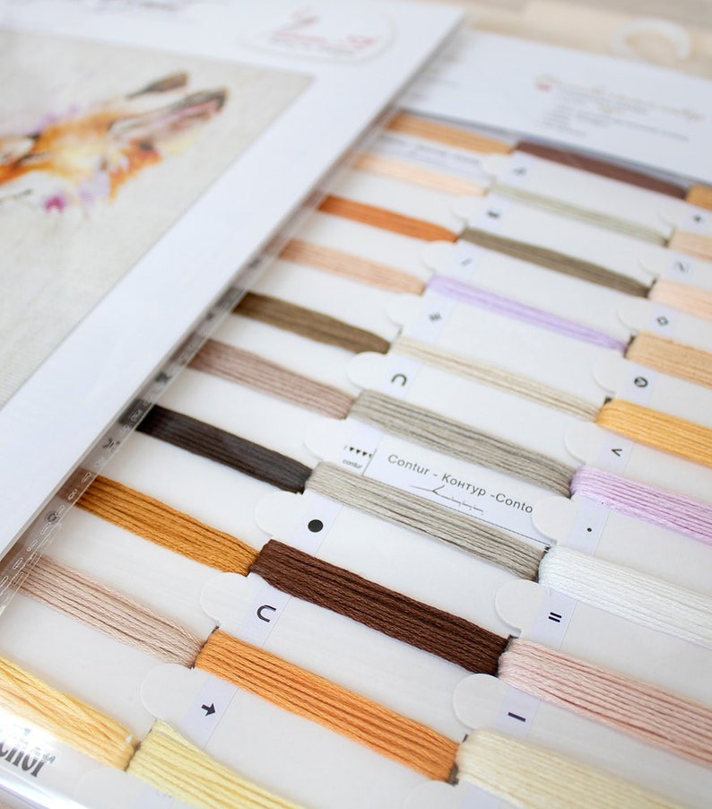 May include: A close-up of a cross-stitch kit with a variety of floss colours, including beige, brown, orange, and white. The floss is organised in labelled sections, with the text "Contour - Контур - Contorno" visible.