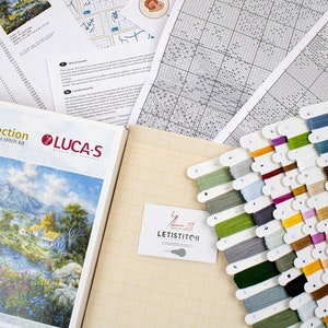 May include: A cross-stitch kit featuring a landscape scene, alongside cross-stitch materials. The kit includes a booklet, fabric, a range of coloured thread spools, and instruction sheets. The brand name "LUCA-S" is visible on the kit.