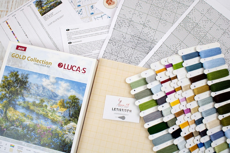May include: A cross-stitch kit featuring a landscape scene, alongside cross-stitch materials. The kit includes a booklet, fabric, a range of coloured thread spools, and instruction sheets. The brand name "LUCA-S" is visible on the kit.