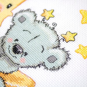 May include: Cross-stitch design of a light blue koala bear embracing a yellow crescent moon. The koala has a sweet expression with pink cheeks and is surrounded by yellow stars on a white background. The stitching is detailed.