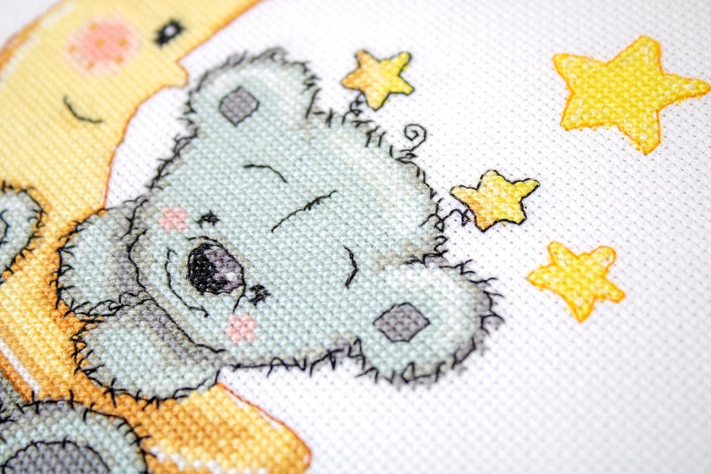 May include: Cross-stitch design of a light blue koala bear embracing a yellow crescent moon. The koala has a sweet expression with pink cheeks and is surrounded by yellow stars on a white background. The stitching is detailed.