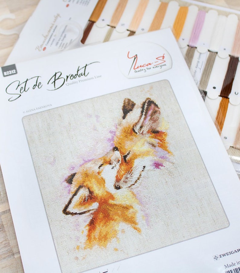May include: Cross stitch pattern featuring two foxes cuddling, with a pink and white background. The pattern is titled "Set de Brodat" and is part of the "Quality Premium Line" by Luca-S.