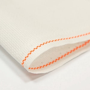 May include: Close-up of white fabric with an orange stitched edge. The fabric has a fine, woven texture.