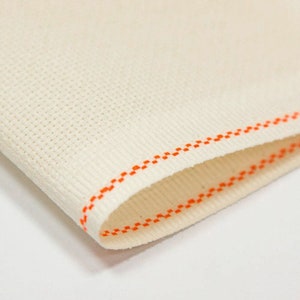 May include: Close-up of a white fabric with an orange stitched border. The fabric has a textured, woven appearance.