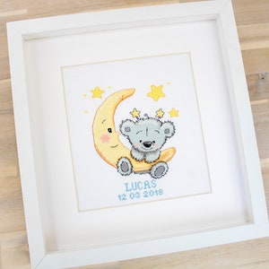 May include: Framed cross-stitch picture. A grey teddy bear sits on a yellow crescent moon, with yellow stars around it. The name "LUCAS" and the date "12 03 2018" are stitched below the bear in a white frame.