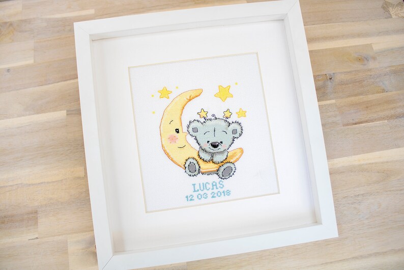 May include: Framed cross-stitch picture. A grey teddy bear sits on a yellow crescent moon, with yellow stars around it. The name "LUCAS" and the date "12 03 2018" are stitched below the bear in a white frame.