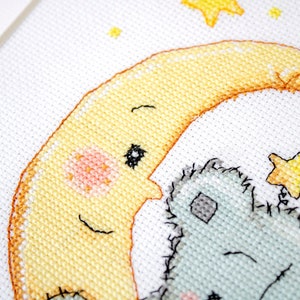 May include: Cross-stitch design showing a smiling yellow moon with a rosy cheek, cuddling a light blue teddy bear. Yellow stars are scattered around the moon and bear. The artwork is on white fabric.
