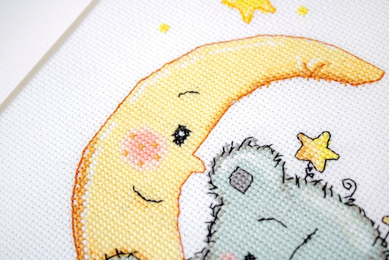 May include: Cross-stitch design showing a smiling yellow moon with a rosy cheek, cuddling a light blue teddy bear. Yellow stars are scattered around the moon and bear. The artwork is on white fabric.