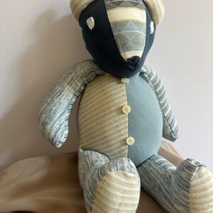 Op de afbeelding: Een blauwe en beige patchwork teddybeer met knoopjesogen en een knoop op zijn borst. De beer zit op een bruin oppervlak.