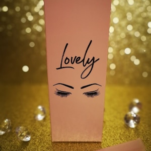 以下が含まれることがあります： ピンクの箱に黒い筆記体で「Lovely」と書かれています。箱には、まつげのある閉じた目の黒いイラストが描かれています。ピンクと白の巻き紙が金色の表面に置かれています。