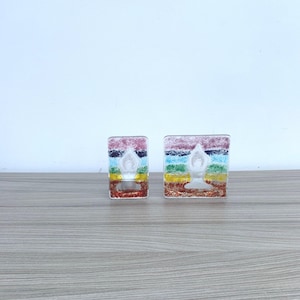 Op de afbeelding: Twee glazen kandelaars met een regenboog chakra ontwerp. Elke houder heeft een witte yoga figuur. De houders zijn rechthoekig en staan op een houten oppervlak.