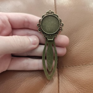 Op de afbeelding: Een antieke bronzen bladwijzer met een decoratief cirkelvormig frame en een lange, slanke, opengewerkte staart. Het frame heeft een sierlijk, wervelend ontwerp. De bladwijzer wordt in een hand gehouden tegen een bruin leren oppervlak.