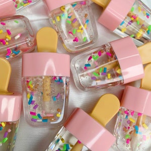 May include: Clear lip gloss containers shaped like ice lollies with pink tops and yellow sticks. The gloss is filled with colourful sprinkles. The containers are arranged on a white surface.