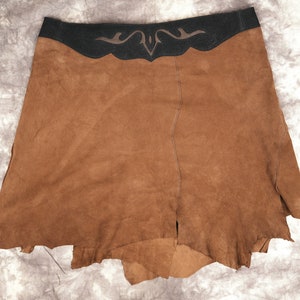 May include: A brown suede skirt with a black leather waistband and a decorative design. The skirt has a raw edge and is fringed.