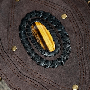 May include: A dark brown suede leather accessory featuring a central oval tiger's eye gemstone. The stone is surrounded by a braided black leather border and secured to the suede base with visible stitching and four small brass-colored metal studs.