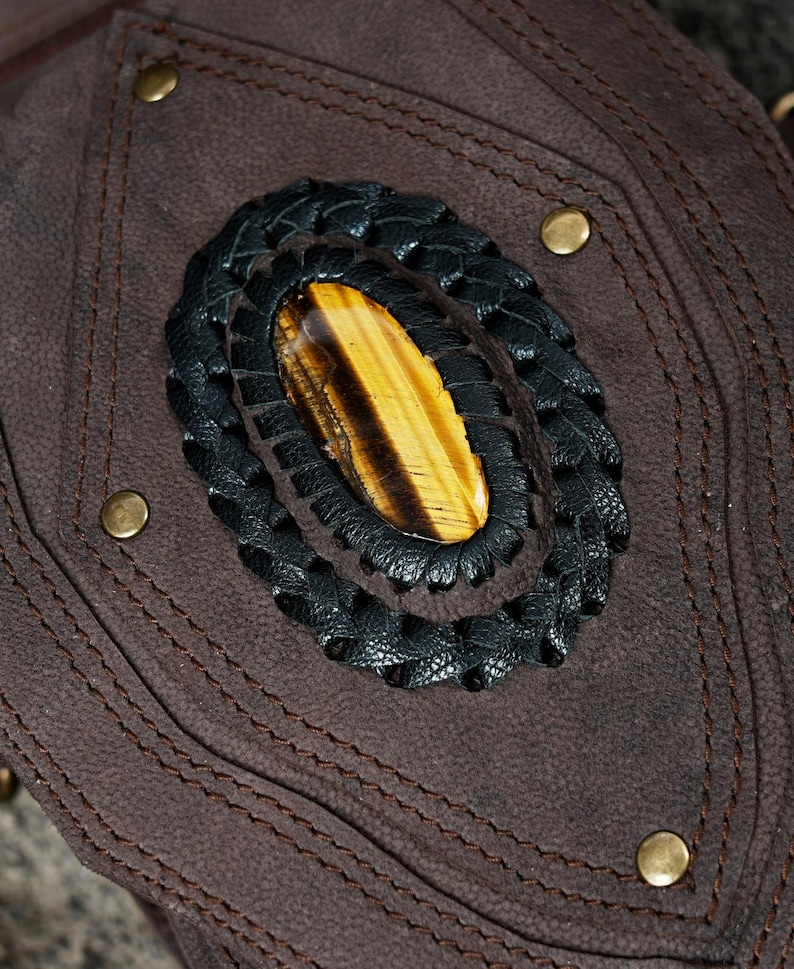 May include: A dark brown suede leather accessory featuring a central oval tiger's eye gemstone. The stone is surrounded by a braided black leather border and secured to the suede base with visible stitching and four small brass-colored metal studs.