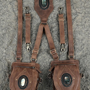 May include: A brown leather harness featuring two attached pouches and adjustable straps with metal hardware. Each pouch and the center back piece are decorated with an oval labradorite gemstone set in a black braided leather border with brass rivets.