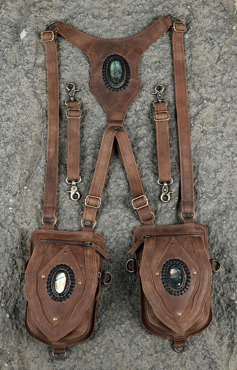 May include: A brown leather harness featuring two attached pouches and adjustable straps with metal hardware. Each pouch and the center back piece are decorated with an oval labradorite gemstone set in a black braided leather border with brass rivets.