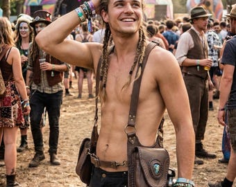 Könnte beinhalten: Ein Mann mit langen geflochtenen Haaren lächelt auf einem Musikfestival. Er trägt eine braune Leder-Umhängetasche mit einem türkisfarbenen Stein, eine schwarze Hose und mehrere Armbänder. Eine Bühne und eine Menschenmenge sind im Hintergrund zu sehen.