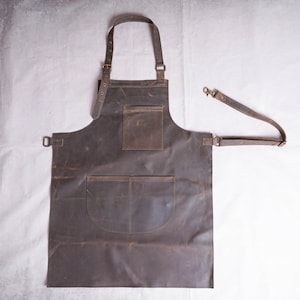 Può includere: Un grembiule in pelle marrone con due grandi tasche e una cinghia regolabile. Il grembiule ha un aspetto usurato e invecchiato.