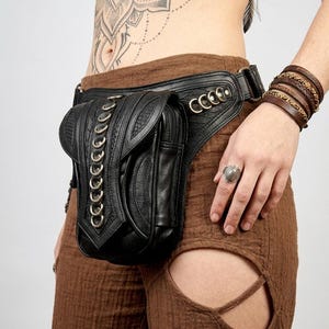Puede incluir: Una riñonera de cuero negro con superficie texturizada, una fila vertical de anillos metálicos plateados en la solapa frontal y una correa ancha con detalles de anillos metálicos adicionales. El bolso cuenta con un cierre de cremallera.