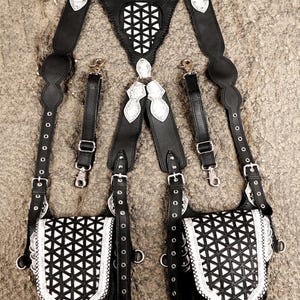 May include: A black leather harness featuring two attached pouches with white geometric triangular cutouts. The harness includes adjustable straps with metal buckles, silver-toned hardware, and two detachable side straps with swivel lobster clasps.