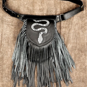 Puede incluir: Un bolso de cintura de cuero negro con flecos largos y una llamativa aplicación de serpiente en la solapa frontal. La serpiente tiene un patrón en blanco y negro que contrasta con el cuero negro liso del bolso y la correa ajustable.