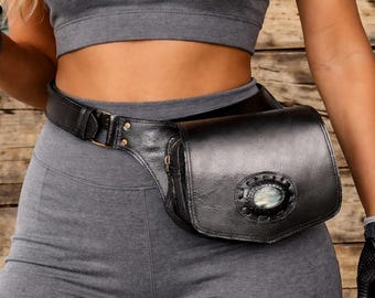 Könnte beinhalten: Schwarze Leder-Bauchtasche mit einem dekorativen ovalen Stein und Nahtdetails. Die Tasche wird mit einem schwarzen Gürtel und einer goldenen Schnalle um die Taille getragen. Das Model trägt graue Sportkleidung.