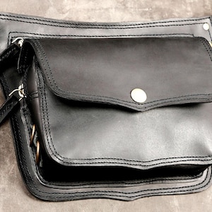 Black leather waist belt bag with front flap closure, metal snap button, antique brass hardware, and multiple zipper pockets for festival fanny pack style.