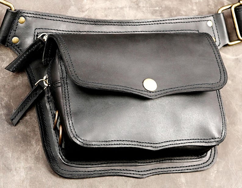 Black leather waist belt bag with front flap closure, metal snap button, antique brass hardware, and multiple zipper pockets for festival fanny pack style.