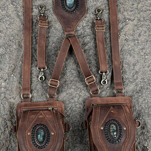 May include: A brown leather harness featuring two attached pouches, each adorned with a central oval gemstone set in a dark braided border. The harness includes adjustable straps with metal buckles and clips, and a back panel with a matching gemstone.