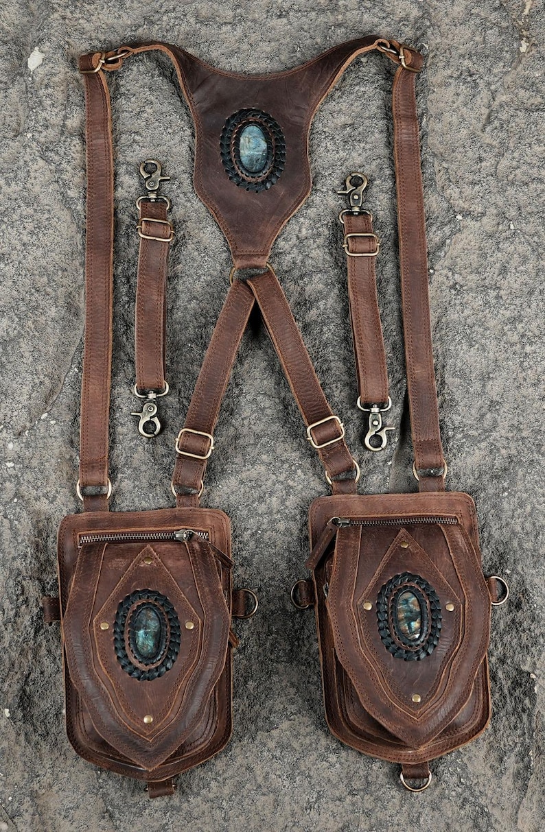May include: A brown leather harness featuring two attached pouches, each adorned with a central oval gemstone set in a dark braided border. The harness includes adjustable straps with metal buckles and clips, and a back panel with a matching gemstone.