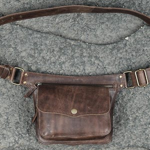 Brown leather utility waist pouch with flap pocket, antique brass hardware, and adjustable strap for hands-free festival wear.