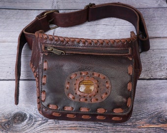 Könnte beinhalten: Braune Leder-Bauchtasche mit Ziernähten und einem dekorativen ovalen Akzent. Die Tasche hat eine Reißverschlusstasche und einen verstellbaren Riemen. Das Leder ist von einem satten, dunklen Braunton, die Nähte sind heller.