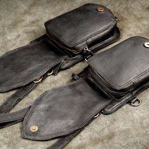 Pair of black leather utility pouches laid flat showing dual zippered compartments and antique brass snap closures.