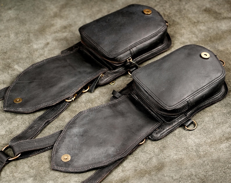 Pair of black leather utility pouches laid flat showing dual zippered compartments and antique brass snap closures.