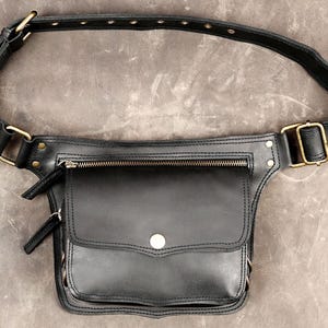 Black handmade leather hip bag featuring antique brass buckle, front zipper pocket, snap flap pouch, and grommet waist strap on grey background. 

Leather Hip Bag, Utility Waist Belt, Festival Fanny Pack, Black Leather Pouch, Pocket Belt, Biker Hip Belt, Travel Waist Bag, Steampunk Belt, Boho Festival Bag, Men Hip Bag, Leather Utility Bag, Handmade Belt Bag, Crossbody Hip Bag