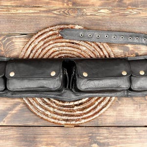 Op de afbeelding: Een zwarte leren riem met vier bevestigde zakken van verschillende groottes, elk beveiligd met een messingkleurige drukknoop. De riem bevat een lange band met meerdere gaatjes en een metalen gesp om rond de taille te dragen.