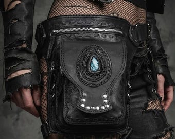 May include: A black leather leg bag featuring a silver zipper closure, a front flap with a teardrop-shaped blue stone inlay, and a row of silver-toned metal studs. The bag includes decorative stitching along the edges and multiple metal rings.
