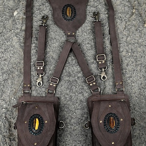 May include: A dark brown leather harness featuring two attached pouches, adjustable straps with metal clips, and three oval tiger eye gemstones set in black braided leather accents. The design includes brass-toned hardware and zippers.