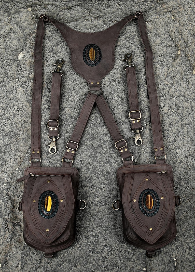 May include: A dark brown leather harness featuring two attached pouches, adjustable straps with metal clips, and three oval tiger eye gemstones set in black braided leather accents. The design includes brass-toned hardware and zippers.