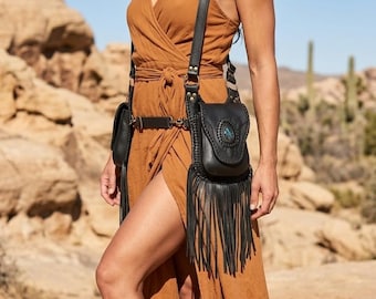 May include: Black leather crossbody bag with fringe and a turquoise stone accent. The bag is worn with a matching belt and a brown dress. The bag has a braided design and a long strap.