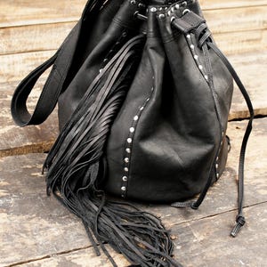 May include: Black leather bucket bag with a long fringe detail and silver studs. The bag has a drawstring closure and a long strap.