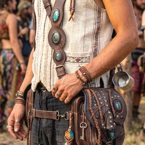 Puede incluir: Un bolso de hombro de cuero marrón con detalles turquesa y tachuelas decorativas. El bolso se lleva con un arnés y cinturón de cuero a juego, y está adornado con borlas coloridas y una llave. La persona lleva un chaleco color crema.