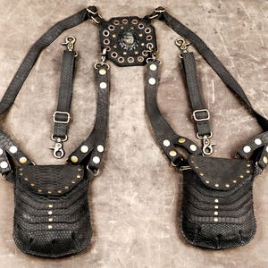 Könnte beinhalten: Ein schwarzes Ledergeschirr mit zwei Taschen in Schlangenlederoptik, messingfarbenen Metallnieten und verstellbaren Riemen. Ein zentrales achteckiges Lederstück mit einem dunklen Steineinsatz verbindet die Schultergurte, die mit Metallclips versehen sind.